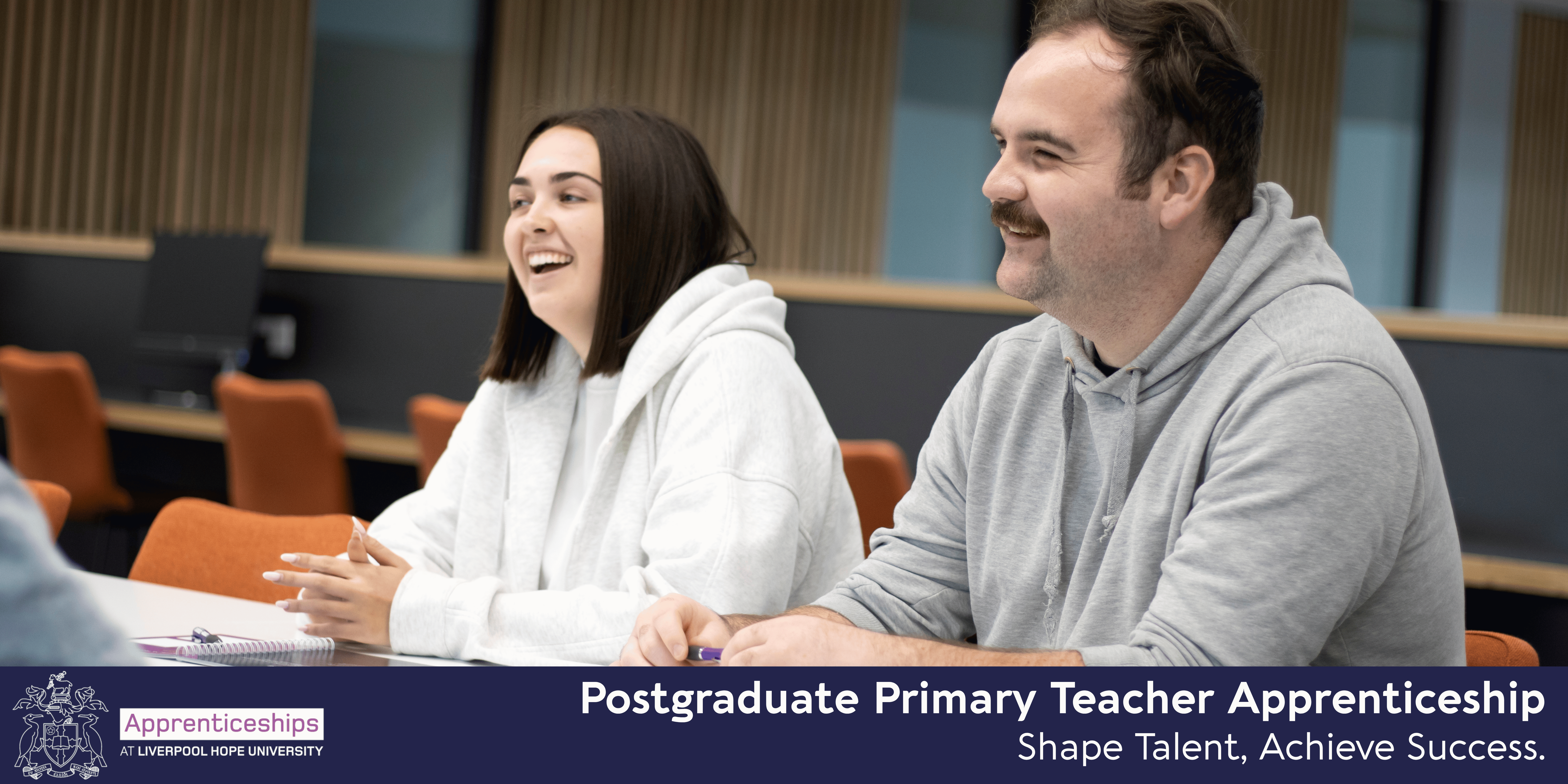 Two students laughing whilst sat at their desk. Banner at the bottom reads Postgraduate Primary Teacher Apprenticeship.